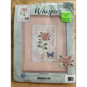 Puede incluir: Un kit de punto de cruz titulado "Delicate Rose" con un diseño floral enmarcado con una rosa rosa y una mariposa azul. El kit incluye hilo de algodón, una aguja e instrucciones. El tamaño de la tela es de 28 x 38 cm. La marca es Dimensions.
