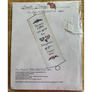 May include: A cross-stitch bell pull kit by Sandi Phipps, featuring the text "Our home is filled with special love." The design includes floral and heart motifs in shades of blue, pink, and purple. The kit includes materials for a 24-count Zweigart linen stitchband.