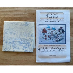 May include: A cream-colored fabric square with a blue bird bath design and a printed pattern. Next to it is a paper with the text "JDR 6023 Bird Bath" and a floral embroidery design. The fabric size is 15" x 15".