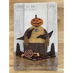 May include: A craft pattern titled "Threads That Bind" featuring a pumpkin-headed figure with crow accents. The figure is wearing a yellow vest and a brown skirt. The text "Crow Catcher" is visible, and the figure is 16 inches tall.