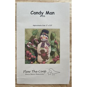 May include: A paper with the text "Candy Man" and "#516" at the top. The image shows a snowman with a scarf and hat, surrounded by greenery and berries. The text "Approximate Size: 3" x 5.5"" is below the title. The bottom of the paper has the text "Flew The Coop" and "PUNCH NEEDLE EMBROIDERY".
