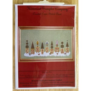 Peut inclure: Une grille de point de croix compté de Historical Sampler Company. Elle présente le mot "CHRISTMAS" avec des arbres et des nichoirs décoratifs. Le motif est encadré d'or sur un fond vert clair avec de la neige. La grille est emballée dans une pochette plastique transparente.