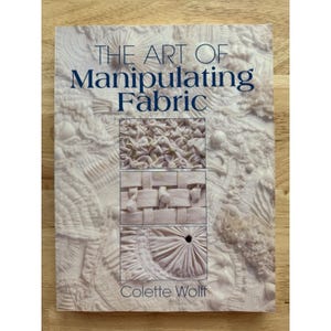May include: A book titled "THE ART OF Manipulating Fabric" with a white cover. The cover features fabric manipulation examples in three rectangular insets. The author's name, Colette Wolff, is at the bottom. The book is on a light brown wooden surface.