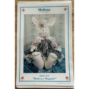 Może przedstawiać: Miękka, biała pluszowa lalka królika o imieniu Melissa, o wysokości około 51 cm, ubrana w kwiecistą sukienkę z koronkowym wykończeniem. Obraz jest wzorem dla lalki, z tekstem "Needle in a Haystack!!".