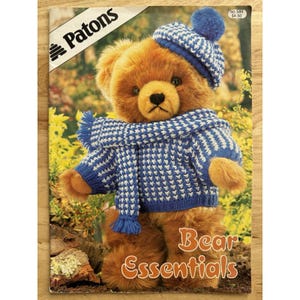 May include: A brown teddy bear wearing a blue and white knit hat, sweater, and scarf. The bear is featured on the cover of a book titled "Bear Essentials" with the Patons logo in the upper left corner.