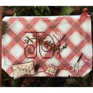 May include: A rectangular needlepoint canvas featuring a pink and white plaid pattern with the word JOY stitched in dark brown, accented by green holly leaves and red berries. Includes two small matching pincushions and a pair of white scissors.