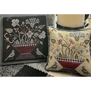 May include: A square cross-stitch sampler on dark fabric and a matching square cross-stitch pincushion on light fabric. Both feature a red vase filled with flowers and two birds, one on each side. The pincushion has a grey ruffled edge.