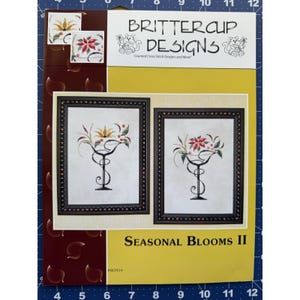 以下が含まれることがあります： Brittercup Designsの「Seasonal Blooms II」というタイトルのクロスステッチキット。このキットには、白黒の2つの額入り花のデザインが特徴です。パッケージは黄色、茶色、白の組み合わせです。「Counted Cross Stitch Designs and More!」というテキストが表示されています。
