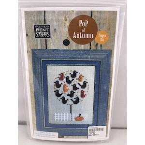 May include: A cross-stitch kit featuring a framed autumn-themed design. The artwork depicts a tree with black birds, orange and red leaves, and a pumpkin. The frame is blue, and the kit is packaged in a clear plastic bag. The text "Pop of Autumn" is visible.