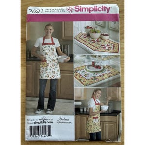 Op de afbeelding: Simplicity naaipatroon 2691 met een schort met kersenprint, een tafelloper en placemats. Maten A, S, M, L en XL inbegrepen. De afbeelding toont een vrouw die het schort draagt in een keuken. Afmetingen in cm.