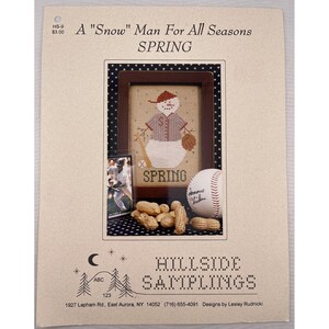 May include: A cross-stitch pattern sheet titled A "Snow" Man For All Seasons SPRING by Hillside Samplings. It features a snowman wearing a baseball cap and jersey, a baseball, a small baseball card, and a pile of peanuts on a dark star background.