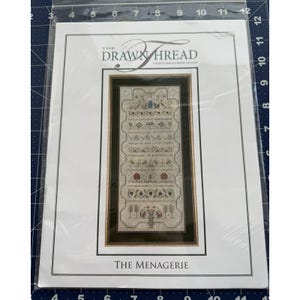 May include: A cross-stitch pattern titled "The Menagerie" by The Drawn Thread. The design features a detailed, framed sampler with various motifs and text. The pattern is packaged in a clear plastic sleeve.