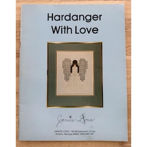 Può includere: Una copertina di libro azzurra con il titolo "Hardanger With Love" in testo nero. La copertina presenta un disegno di angelo a punto croce incorniciato. La firma dell'artista, "Janice Love", è in basso.