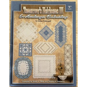 Op de afbeelding: Een boekomslag getiteld "Tomorrow's Heirlooms III in Hardanger Embroidery" met diverse geborduurde ontwerpen in blauw en wit. De omslag bevat tekst van Earlene Dobberpuhl en samengesteld door Susan L. Meier en Rosalyn K. Watnemo.