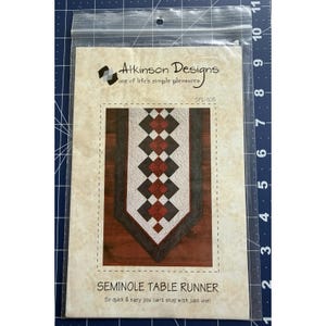 May include: A sewing pattern for a Seminole table runner. The pattern features a geometric design in red, white, and brown. The package includes the Atkinson Designs logo and the text "So quick & easy you can't stop with just one!"