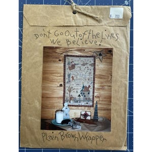 May include: A brown paper package with the text "Don't Go Out of the Lines We Believe!" and "Plain Brown Wrapper". Inside is a framed Christmas-themed cross-stitch with a wooden background. A small price tag is visible.