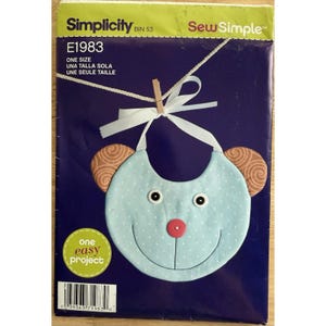 May include: A Simplicity sewing pattern for a baby bib in the shape of a bear's face. The bib is light blue with white polka dots, brown ears, and a red nose. The pattern is labelled E1983 and includes the text "one easy project".