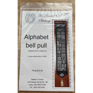 Peut inclure: Un modèle de point de croix pour un tire-cloche alphabet, avec un fond noir et des motifs géométriques en blanc et or. L'emballage comprend le texte "Alphabet bell pull" et "The Framed Owl Stitchery".