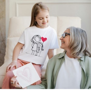 May include: A white t-shirt with a black and white illustration of two stick figures holding a red heart balloon. The figures are a mother and daughter. A young girl is wearing the shirt and smiling at an older woman.
