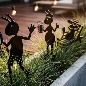 Könnte beinhalten: Schwarze Metall-Gartenstecker, die Cartoon-Ameisen bei Freizeitaktivitäten darstellen. Eine Ameise winkt, eine andere hält ein Bier und eine dritte entspannt sich in einem Stuhl, ebenfalls mit einem Bier. Die Stecker sind in einem Beet aus grünem Gras platziert.