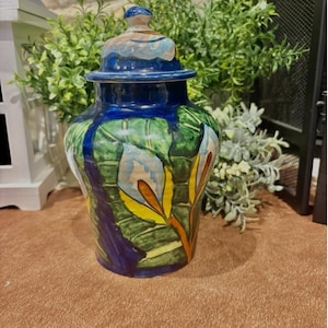 May include: A blue and green ceramic jar with a lid. The jar is decorated with a floral design in shades of yellow, orange, white, and blue.