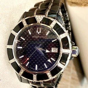 Może przedstawiać: Czarny ceramiczny zegarek z kopertą wysadzaną diamentami i niebieską tarczą z włókna węglowego. Tarcza zegarka ma srebrne wskazówki, białe indeksy godzinowe i datownik w okienku na godzinie 3. Zegarek jest oznaczony jako "Accutron" i "Swiss Made".