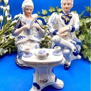 Könnte beinhalten: Eine Porzellanfiguren-Garnitur mit einem Paar, das an einem Tisch mit einer Teekanne und zwei Tassen sitzt. Die Figuren sind in Blau und Weiß mit goldenen Akzenten bemalt.