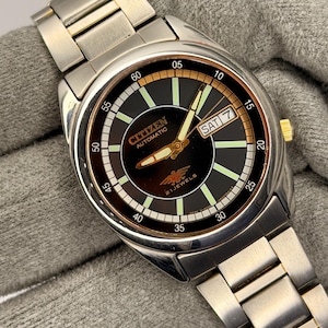 Peut inclure: Une montre-bracelet automatique Citizen en acier inoxydable avec un cadran noir. Le cadran affiche le jour et la date, avec des aiguilles et des marqueurs lumineux. Le bracelet est en métal argenté et la couronne dorée.
