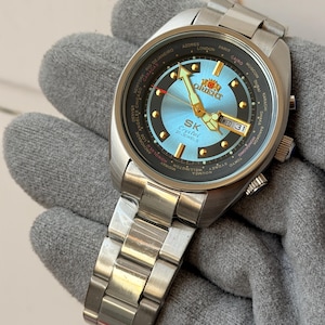 Peut inclure: Une montre-bracelet Orient SK Crystal argentée avec un cadran bleu clair et un anneau extérieur noir affichant les villes du monde. La montre comporte un affichage de la date et le texte "SK Crystal 21 Jewels". La montre est sur un bracelet en métal argenté.