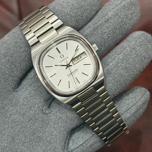 Pode incluir: Um relógio de pulso automático Omega Seamaster prateado com mostrador retangular. O mostrador é prateado com marcadores de hora e ponteiros pretos. O dia e a data são exibidos em uma pequena janela. A pulseira é de metal prateado.