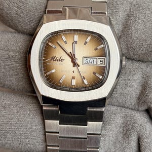 Puede incluir: Un reloj de pulsera automático Mido Multi-Star vintage. El reloj presenta una caja y brazalete plateados, una esfera marrón y una pantalla de día y fecha. La esfera del reloj tiene agujas e indicadores plateados. El reloj se muestra sobre una superficie gris.