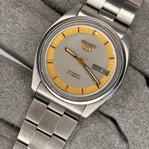 May include: A silver Seiko 5 automatic watch with a gray face and gold accents. The watch features a day and date display and a silver metal band. The watch face has the text "SEIKO 5 AUTOMATIC 17 JEWELS".