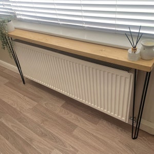 May include: A wooden console table with a light-coloured top and black hairpin legs. The table is positioned in front of a white radiator. A small potted plant and a diffuser are on the table. The background includes a window with blinds.