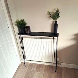 May include: A black console table with a wooden top and hairpin legs. The table is decorated with a potted plant, a candle, and a vase with greenery.