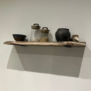 May include: A wooden shelf with a rustic finish, featuring a black ceramic pot, three glass jars with lids, and a blue and brown ceramic bowl. The shelf is mounted on a white wall with black metal brackets.