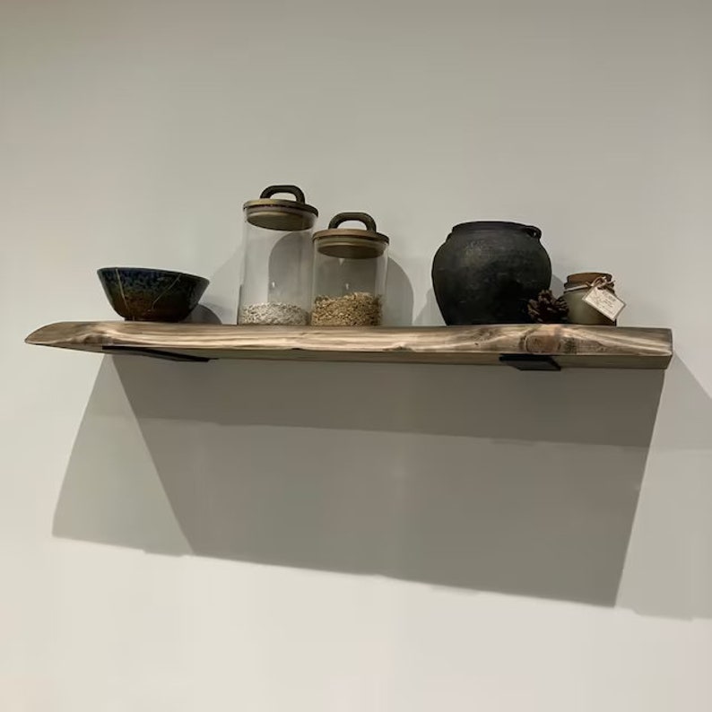 May include: A wooden shelf with a rustic finish, featuring a black ceramic pot, three glass jars with lids, and a blue and brown ceramic bowl. The shelf is mounted on a white wall with black metal brackets.