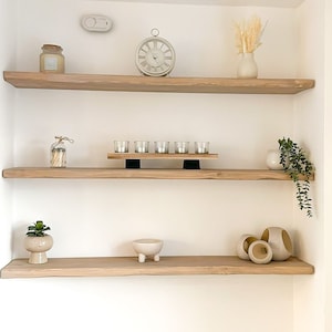 May include: Three wooden shelves with various decorative items, including a clock, candles, a vase with dried flowers, a potted plant, and a small ceramic bowl. The shelves are mounted on a white wall.