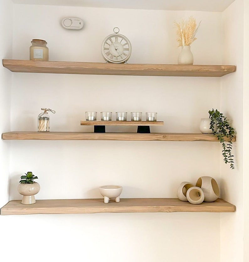 May include: Three wooden shelves with various decorative items, including a clock, candles, a vase with dried flowers, a potted plant, and a small ceramic bowl. The shelves are mounted on a white wall.