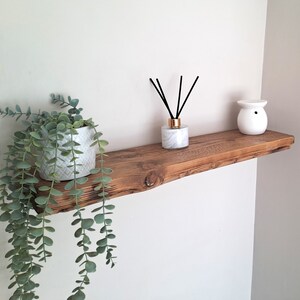 May include: A rustic wooden shelf with a white ceramic diffuser, a plant in a white pot, and black diffuser sticks. The shelf is mounted on a white wall.