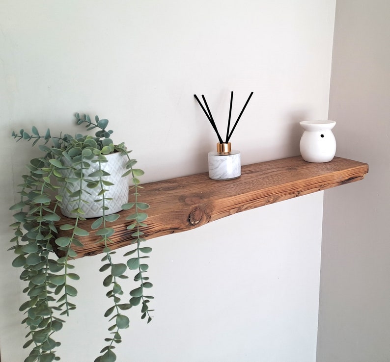 May include: A rustic wooden shelf with a white ceramic diffuser, a plant in a white pot, and black diffuser sticks. The shelf is mounted on a white wall.