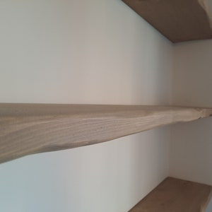 May include: Close-up of a light brown wooden shelf with a natural, rough-hewn edge. The shelf is mounted on a white wall.