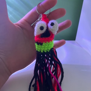 May include: A handmade crochet keychain shaped like a cartoon squid. The squid has large white eyes, a red head, a green midsection, and black tentacles with red and pink accents. The keychain is attached to a silver metal ring.