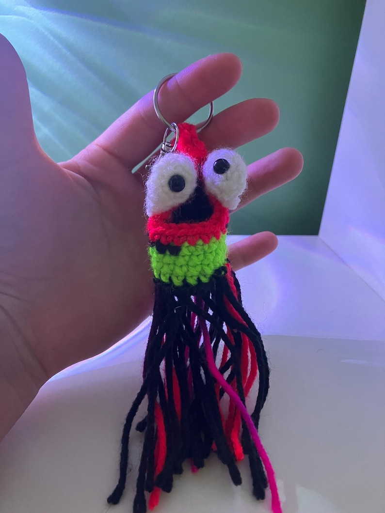 K&ouml;nnte beinhalten: Ein handgefertigter Schl&uuml;sselanh&auml;nger aus H&auml;kelarbeit in Form eines Cartoon-Tintenfischs. Der Tintenfisch hat gro&szlig;e wei&szlig;e Augen, einen roten Kopf, einen gr&uuml;nen Mittelteil und schwarze Tentakel mit roten und rosa Akzenten. Er ist an einem silbernen Metallring befestigt.