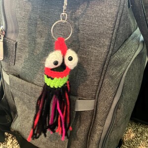 K&ouml;nnte beinhalten: Ein handgefertigter H&auml;kel-Schl&uuml;sselanh&auml;nger mit einem skurrilen Tintenfisch-Design. Der Tintenfisch hat einen roten Kopf, gro&szlig;e wei&szlig;e Augen mit schwarzen Pupillen und einen gr&uuml;nen und schwarzen K&ouml;rper. Die Tentakel sind schwarz, rosa und rot. Der Schl&uuml;sselanh&auml;nger ist an einem silbernen Metallring und Verschluss befestigt.