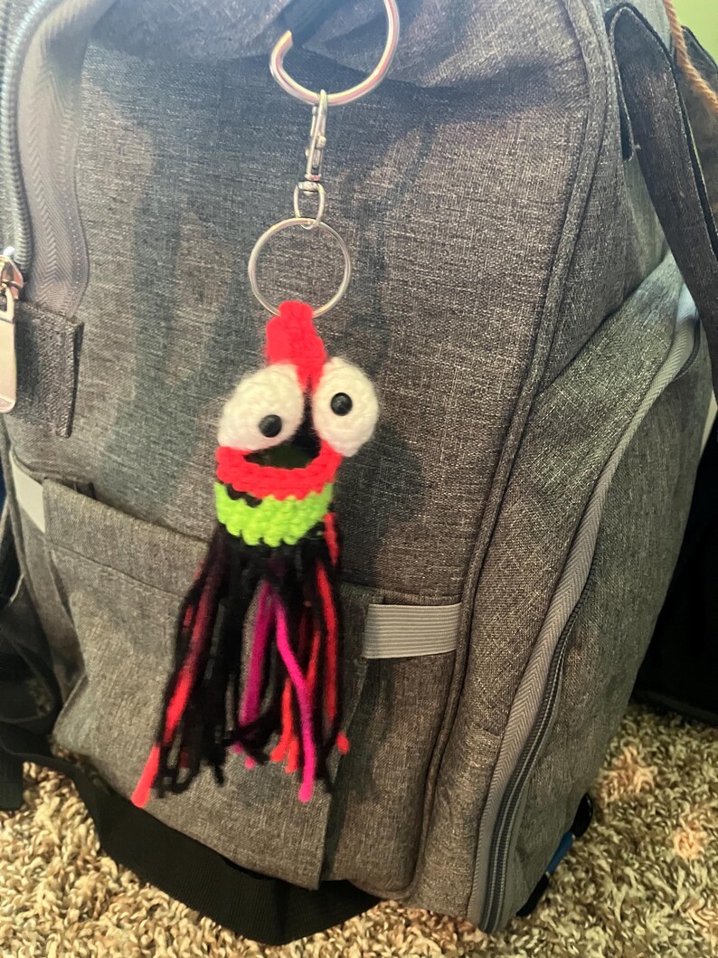 K&ouml;nnte beinhalten: Ein handgefertigter H&auml;kel-Schl&uuml;sselanh&auml;nger mit einem skurrilen Tintenfisch-Design. Der Tintenfisch hat einen roten Kopf, gro&szlig;e wei&szlig;e Augen mit schwarzen Pupillen und einen gr&uuml;nen und schwarzen K&ouml;rper. Die Tentakel sind schwarz, rosa und rot. Der Schl&uuml;sselanh&auml;nger ist an einem silbernen Metallring und Verschluss befestigt.