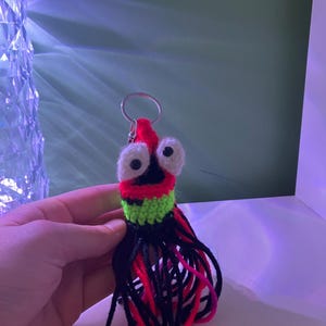 K&ouml;nnte beinhalten: Ein handgefertigter H&auml;kel-Schl&uuml;sselanh&auml;nger mit einem skurrilen Kreaturen-Design. Der Schl&uuml;sselanh&auml;nger hat einen roten Kopf mit gro&szlig;en wei&szlig;en Augen, einen gr&uuml;nen und schwarzen K&ouml;rper und einen schwarzen, roten und pinkfarbenen Fransen. Ein silberner Schl&uuml;sselring ist befestigt.