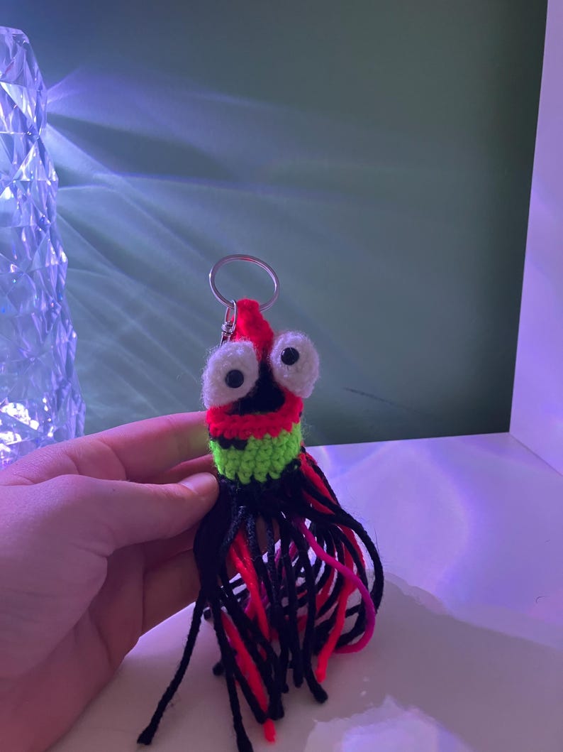 K&ouml;nnte beinhalten: Ein handgefertigter H&auml;kel-Schl&uuml;sselanh&auml;nger mit einem skurrilen Kreaturen-Design. Der Schl&uuml;sselanh&auml;nger hat einen roten Kopf mit gro&szlig;en wei&szlig;en Augen, einen gr&uuml;nen und schwarzen K&ouml;rper und einen schwarzen, roten und pinkfarbenen Fransen. Ein silberner Schl&uuml;sselring ist befestigt.