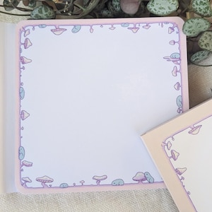 Può includere: Un blocco note quadrato con un bordo rosa chiaro con un motivo ripetuto di funghi dei cartoni animati in vari colori. Il blocco note è aperto, rivelando pagine bianche vuote. È visibile un blocco note più piccolo con un design simile.