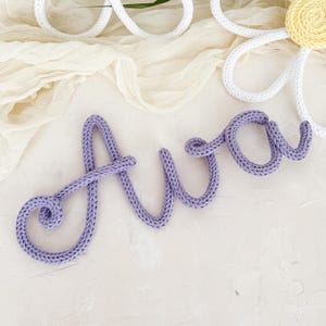 Könnte beinhalten: Zwei handgefertigte Gänseblümchen-Dekorationen mit weißen Blütenblättern und gelben Zentren. Ein lavendelfarbener gestrickter Name, "Ava", ist ebenfalls vorhanden. Die Gänseblümchen und der Name sind auf einem cremefarbenen Stoff mit grünen Blättern im Hintergrund platziert.