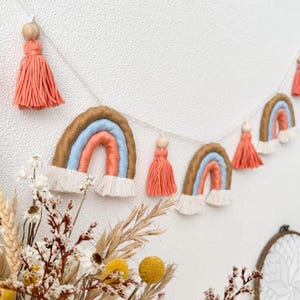 Op de afbeelding: Een guirlande van vijf regenboogkleurige macramé regenbogen met perzikkleurige kwastjes die aan een wit touw hangen. De regenbogen zijn gemaakt van bruin, blauw en perzikkleurig garen.